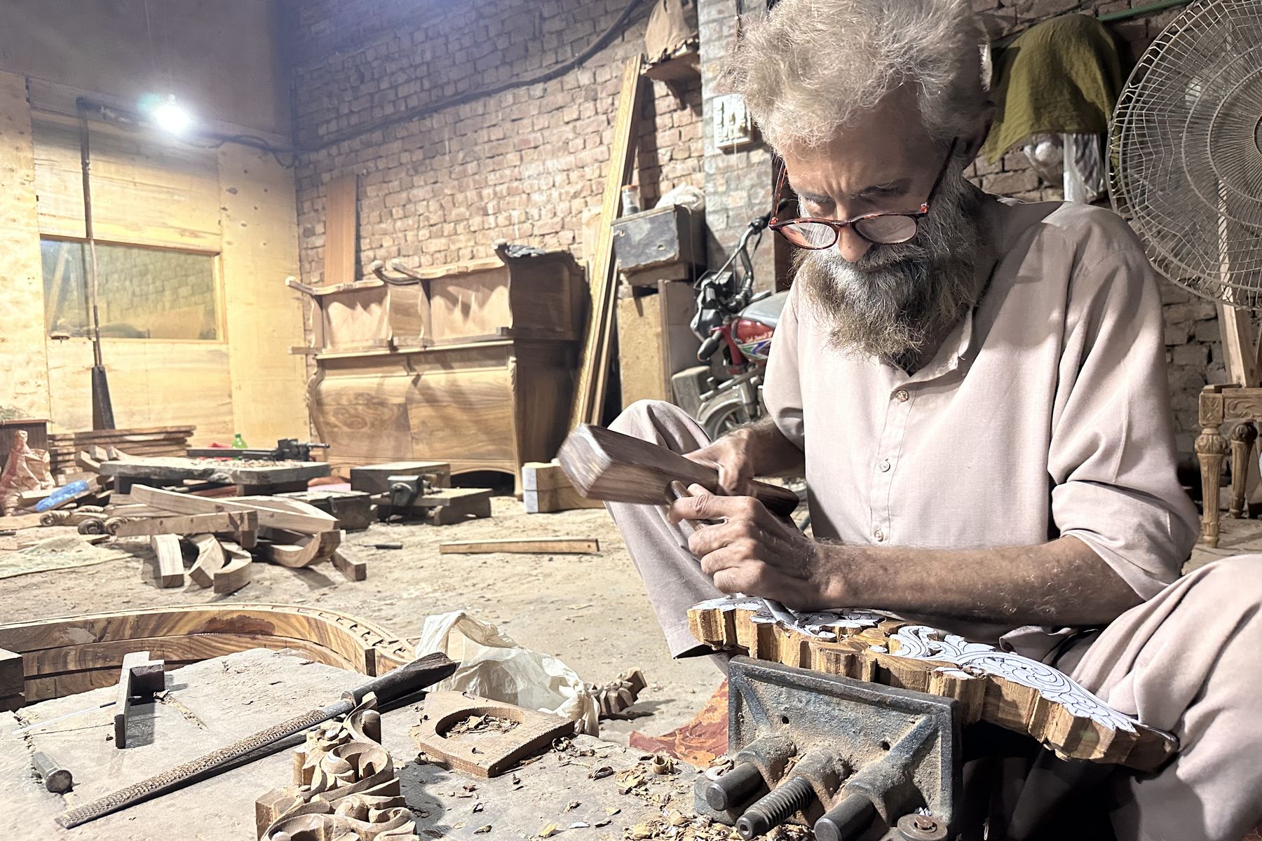 A senior craftsman carving an ornamental wooden panel held in a vice in the Chiniot workshop