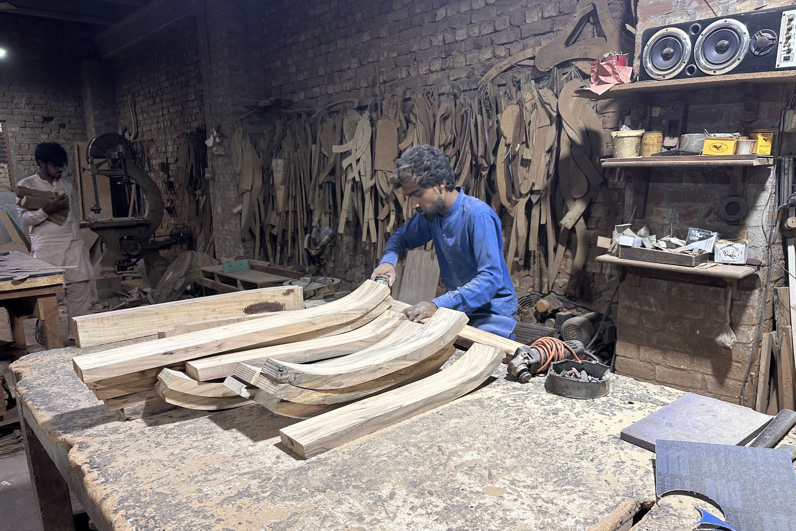 A craftsman shaping curved solid wood frame parts beneath a wall of hanging wooden templates in the Chiniot workshop